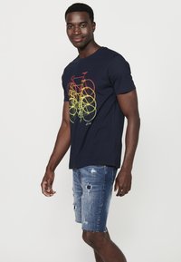 Camiseta azul marino con un gráfico de bicicleta multicolor en rojo y amarillo. Combinada con pantalones cortos de mezclilla desgastados. Mangas cortas, cuello redondo.