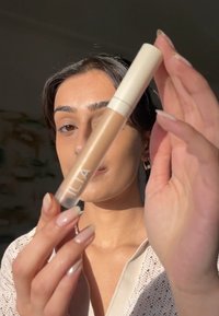 Korektor ILIA w przezroczystej tubce, zawierający beżową płynną formułę, z białą nakrętką w kształcie aplikatora. Ręce z nude lakierem do paznokci. Delikatne oświetlenie.