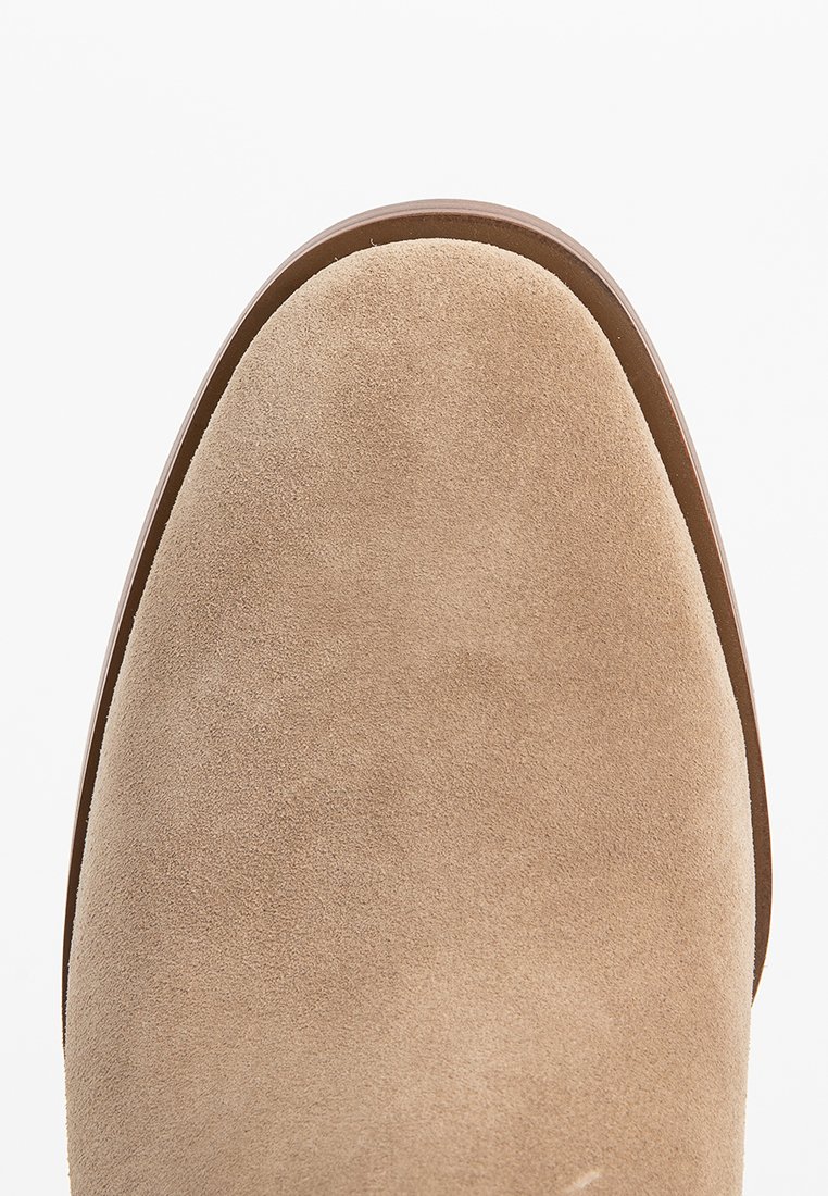 Gros plan de l'orteil d'une chaussure en daim beige avec un bord de semelle lisse marron sur fond blanc.