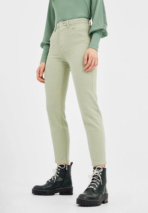 Personne portant un pantalon taille haute vert clair, un haut vert côtelé manches longues et des bottes de combat noires avec des lacets blancs.