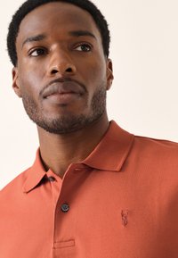 Poloshirt in roestkleur met een gestructureerde stof, twee knopen bij de kraag en een klein geborduurd logo op de linkerborst.