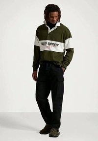 Polo de manga comprida verde-oliva com riscas brancas e texto "POLO SPORT". Calças cargo pretas; sapatos com sola de borracha. Modelo em postura descontraída.