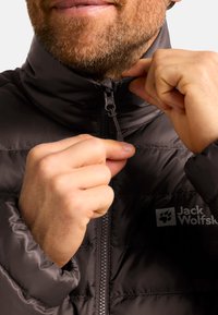 Black puffer jacket with a high collar and zipper. Features a smooth, glossy texture and a logo on the left side. Hands adjusting the zipper.