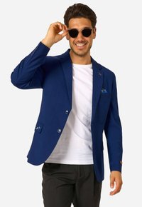 Vīrietis smaidot pielāgo melnas apaļas saulesbrilles, tērpies z blue blāzerī, baltajā krekliņā un melnās biksēs uz vienkārša fona.