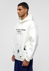 Witte hoodie met een voorzak, voorzien van zwart en blauwe verfspatten. Zwarte tekst op de borst luidt "REVOLUTIONEER ART DESIGN PROCESSEN."