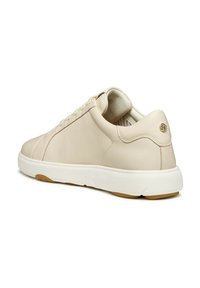 Zapatilla de cuero beige con forma redondeada, que presenta una suela de goma blanca y un logotipo dorado en el talón, mostrando detalles de costura.
