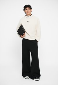Krämfärgad sweatshirt med en åtsittande halsringning och logotyp, i kombination med svarta vida byxor och klumpiga svartvita sneakers. Håller en svart väska.