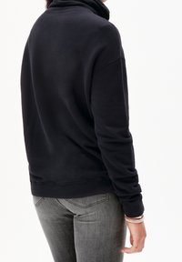 Osoba v černém topu s dlouhým rukávem a šedých džínách, zobrazena zezadu s bílou gumičkou na zápěstí.