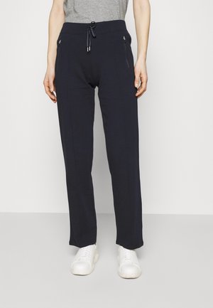 Donna che indossa pantaloni navy con coulisse e tasche con cerniera, e sneakers bianche, in piedi davanti a uno sfondo chiaro e uniforme.