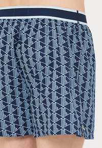 Şorturi de înot navy cu un model geometric de culoare albastru deschis. Prezintă o talie elastică cu un accent albastru în contrast și detalii pe cusătura laterală.