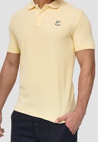 Mies, ktorý má na sebe svetložlté polo tričko s krátkym rukávom a malým čiernym logom na ľavej hrudi, v kombinácii s tmavými nohavicami, ruku má v vrecku.