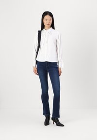 Chemisier blanc avec un nœud au cou, boutons à l'avant et manches longues, associé à un jean bleu foncé. Porté avec des bottines noires et un sac noir.