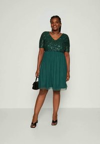 Robe verte à sequins avec des manches courtes, décolleté en V et une jupe fluide et transparente. Portée avec des chaussures noires et un petit sac à main noir.