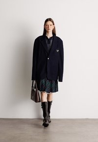 Υπερμεγέθης ναυτικό σακάκι φορεμένο πάνω από ένα σκούρο πουκάμισο και καρό φούστα, συνδυασμένο με μαύρες μπότες μέχρι το γόνατο και καφέ τσάντα.