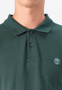 Tummanvihreä polo-paita, joka on valmistettu teksturoidusta kankaasta. Paidan muotoilu sisältää klassisen kauluksen, kaksi mustaa nappia ja pienen vaaleanvihreän brodeeratun logon.