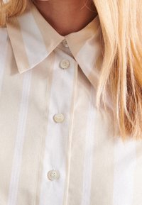 Beige and white striped button-up shirt with pointed collar and long blonde hair partially visible on the right side.