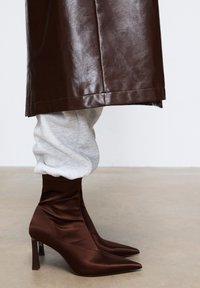 Stivali alla caviglia in satin marrone con punta appuntita e tacco alto, caratterizzati da un design arricciato alla caviglia, abbinati a pantaloni della tuta grigi chiari.