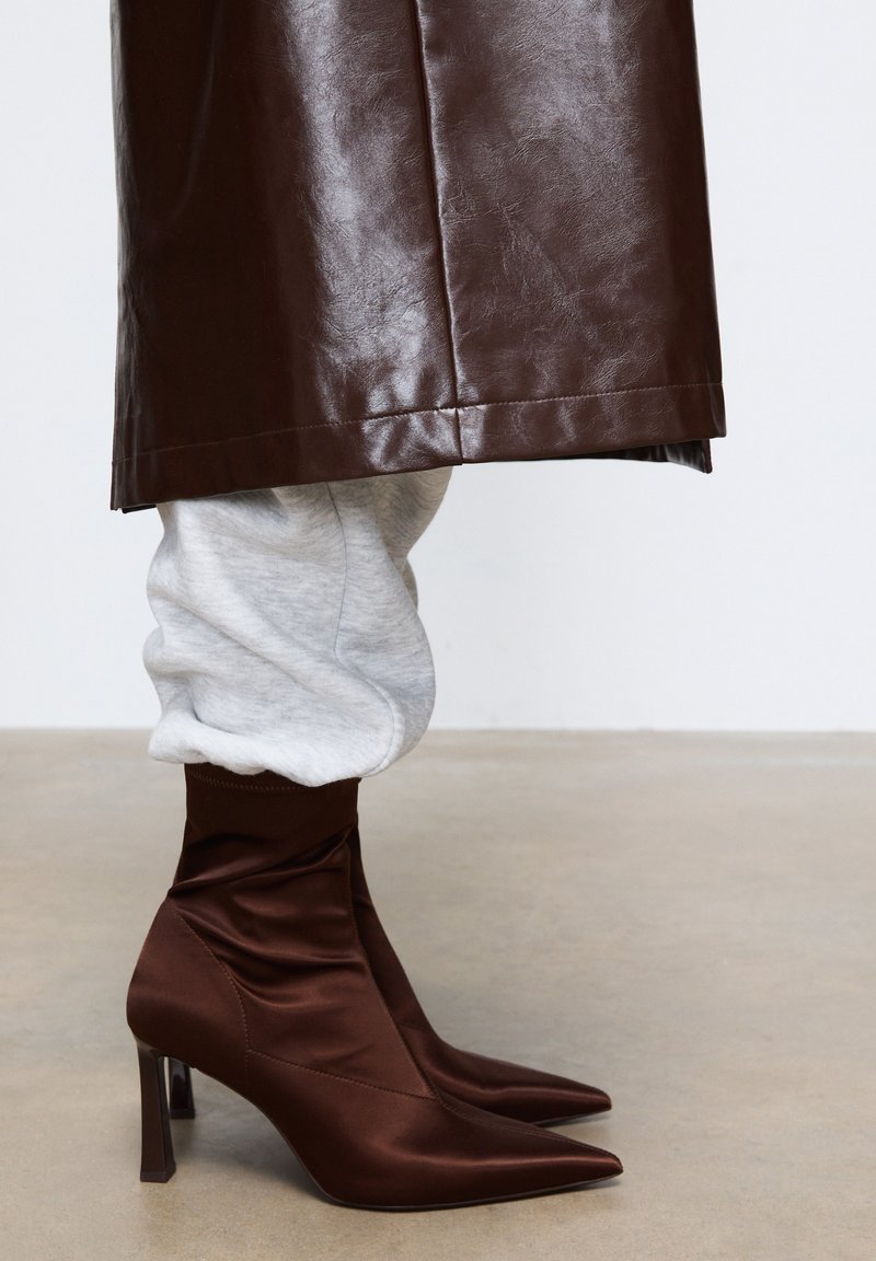 Stivali alla caviglia in satin marrone con punta appuntita e tacco alto, caratterizzati da un design arricciato alla caviglia, abbinati a pantaloni della tuta grigi chiari.