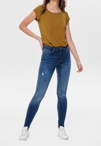 Senapsgul kortärmad topp tillsammans med åtsittande blå denimjeans som visar lätt slitage. Bärs med vita sneakers på en neutral bakgrund.