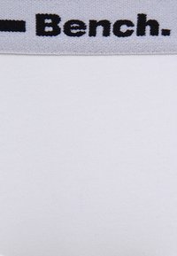 Biely textil s hladkou štruktúrou, ktorý má na vrchu šedý pás, na ktorom je v tučných čiernych písmenách napísané slovo "Bench".