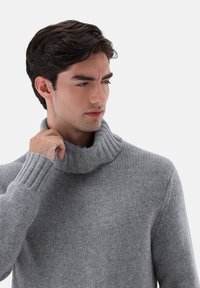 Grau gestrickter Pullover mit hohem Rippenkragen und langen Ärmeln, der ein strukturiertes Muster und eine lockere Passform aufweist.