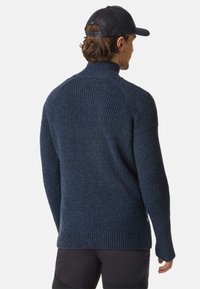 Mørkeblå strikket sweater med høj krave, ribbet tekstur og lange ærmer. Har et logo-accent ved den nederste side. Blødt, varmt materiale.