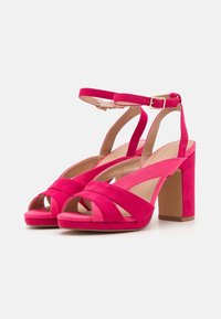 Sandales à talons hauts en daim rose avec des brides croisées, design à bout ouvert, sangle de cheville avec boucle, et un talon épais. Texture lisse sur l'ensemble.