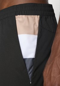 Svart sportshorts har en sidoventil som visar ett lager av tyg i beige, vitt och grått med ett strukturerat mönster. Elastisk midja ingår.