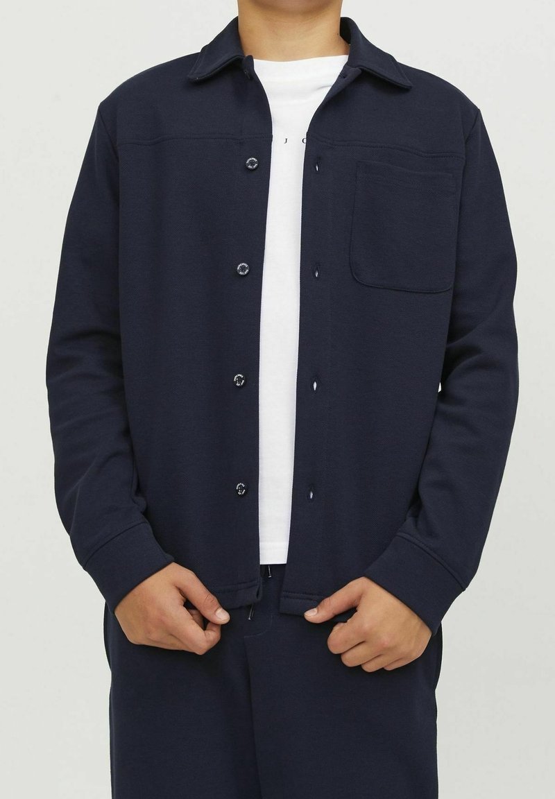 Veste boutonnée bleu marine avec un col, une poche poitrine et un tissu texturé. Le mannequin porte un T-shirt blanc en dessous.