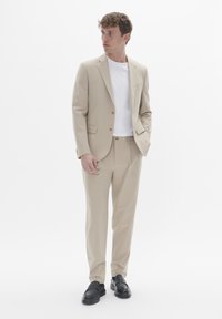 Costume beige avec un blazer à un bouton, un pantalon assorti, un t-shirt blanc et des mocassins noirs. Tissu lisse avec des accents de boutons.