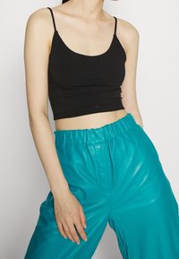 Femme portant un crop top noir à fines bretelles et un pantalon en cuir bleu sarcelle taille haute avec une ceinture élastique.