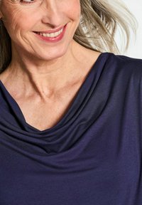 Marineblauwe stof met drapeer effect, voorzien van een kettinghalslijn, zachte textuur en een gladde afwerking met een subtiele glans, waarbij het sleutelbeen zichtbaar is.
