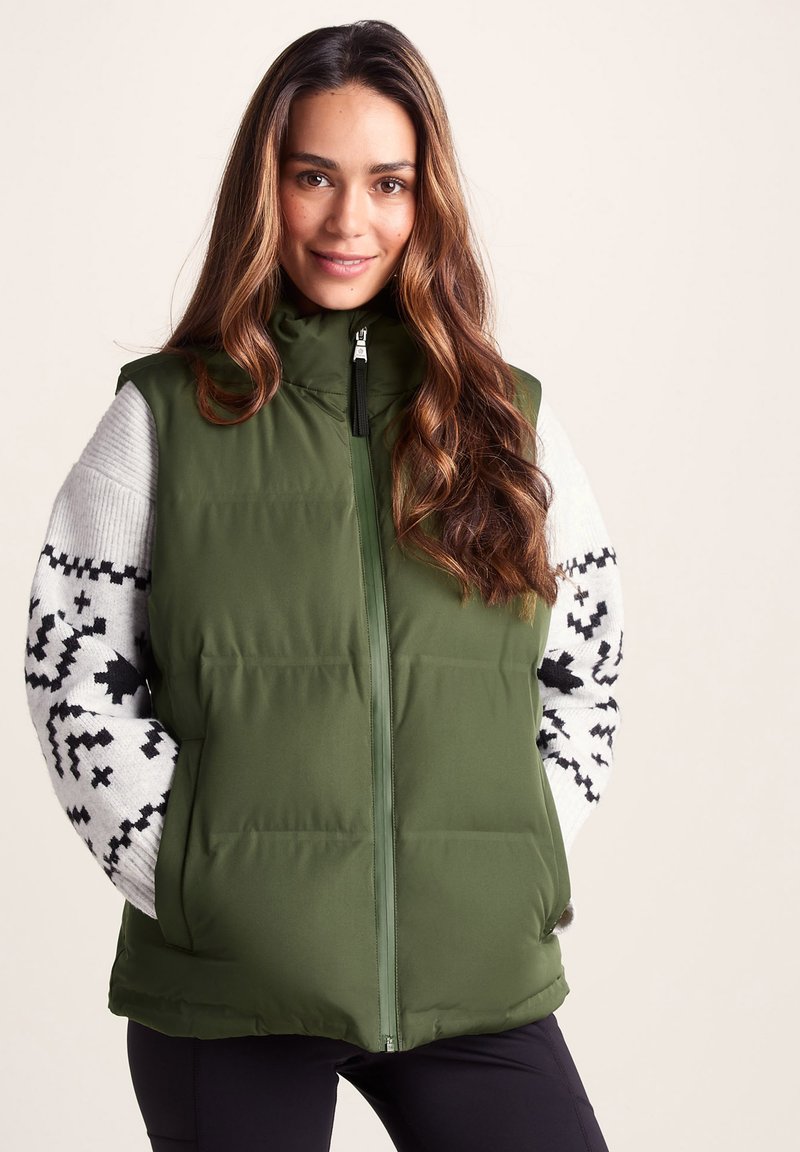 Olive green puffer vest with a high collar, front zipper, and black hardware; paired with a patterned sweater featuring black and white designs.