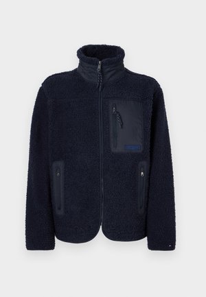 Veste polaire bleu marine à fermeture éclair intégrale, col montant, deux poches latérales zippées et une poche poitrine zippée avec des tirettes en tissu.