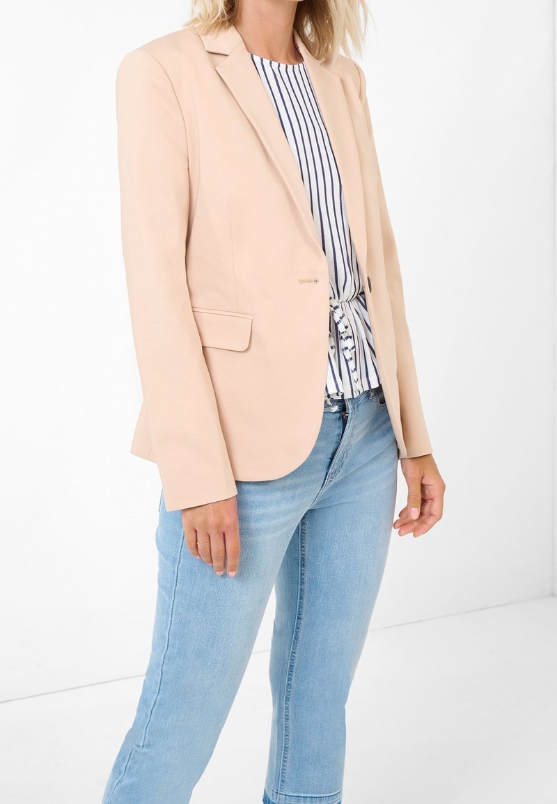 Femme portant un blazer beige sur un chemisier blanc et bleu rayé noué, avec un jean bleu clair, debout devant un fond blanc.