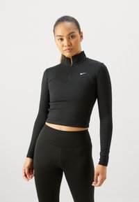 Musta pitkähihainen crop-top, jossa on vetoketjukaulus ja Nike-logo rinnassa, valmistettu venyvästä materiaalista, yhdistettynä korkeavyötäröisiin mustiin leggings-housuihin.