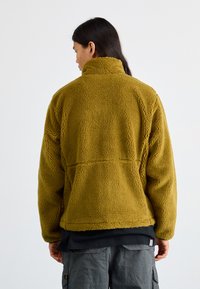 Veste en polaire verte olive avec une surface texturée, col montant et coupe décontractée. Présente une couture dans le dos pour un détail de design.