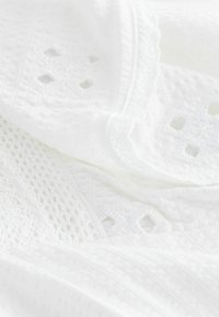 Biała, fakturowana tkanina z misterną koronką i otwartymi wzorami w diamenty. Miękka i lekka, z finezyjnymi detalami wzdłuż brzegów.