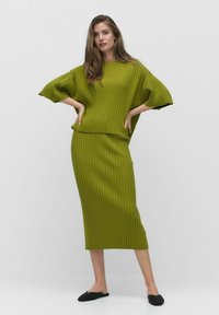 Vihreä ribattu neuleasu, jossa on väljä yläosa leveillä hihoilla ja tiukka midi-hame. Mustat liukuavat kengät täydentävät ilmeen.