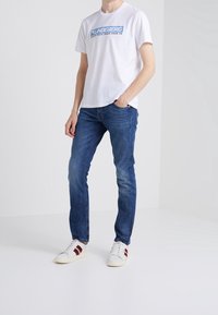 Vit t-shirt med blå grafisk tryck, blå denimjeans med blekningsdetaljer och vita sneakers med röda ränder.