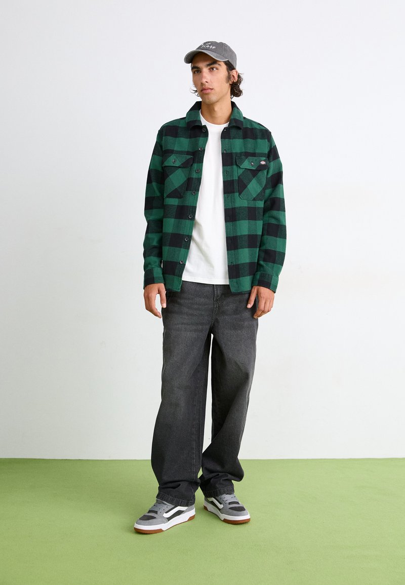 Chemise en flanelle à carreaux verts et noirs avec deux poches poitrine, associée à un t-shirt blanc, un jean sombre et des baskets grises sur une surface verte.