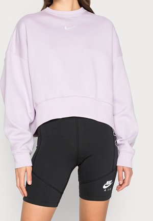 Kobieta ubrane w jasnofioletową krótką bluzę i czarne spodenki rowerowe Nike Air, stoi z rękami swobodnie opuszczonymi wzdłuż ciała.