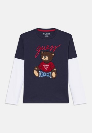 Navyblaues langärmliges T-Shirt, das ein Grafikdesign eines Braunbären mit rotem Pullover und Jeanshose zeigt, mit dem aufgestickten Wort 'guess' in Rot darüber.