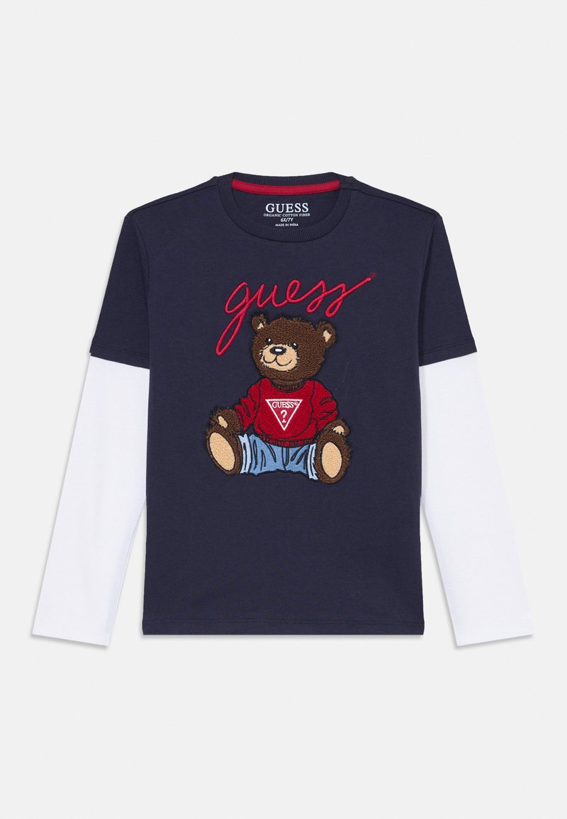 Tamsiai mėlyna ilgomis rankovėmis marškinėliai su rudo meškos grafika, vilkinčia raudoną megztinį ir džinsus, o viršuje raudonai išsiuvinėtas tekstas „guess“.