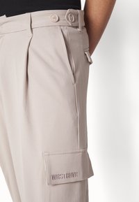 Ljust beige cargobyxor med dubbelknäppning i midjan, veck, sidoficka och broderad detalj "WRSTBHVR" på cargofickan.