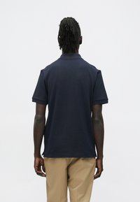 Hátratekintő férfi fonott hajjal, sötétkék rövid ujjú galléros pólót és bézs nadrágot visel, egyszerű háttér előtt.