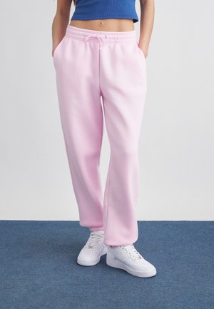 Kobieta ubrana w jasnoróżowe spodnie dresowe z troczkiem w pasie, białe trampki i niebieski krótki top, stoi na niebieskim dywanie.