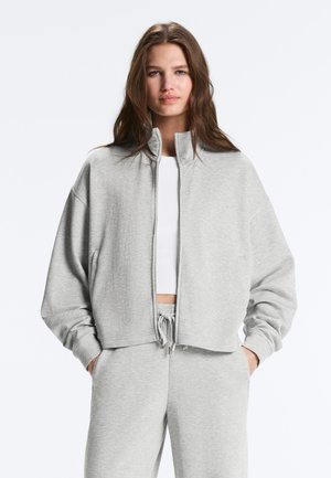 Fiatal nő hosszú barna hajjal, világosszürke cipzáras pulóvert és hozzá illő melegítőnadrágot visel, kezét zsebében tartja, semleges háttér előtt.