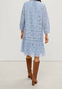 Femme portant une robe en dentelle florale bleu clair à manches longues et à ourlet évasé, associée à des bottes en daim marron à hauteur de genou avec talons, de dos.