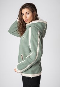 Groene fleecejas met capuchon, crème accenten op de mouwen, ritszakken en een rond logo op de borst. Gestructureerd, zacht materiaal.